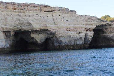 Güney Portekiz 'deki Algarve kıyısı boyunca uzanan kayalık manzaralı bir uçurum, sıcak güneş ışığıyla yıkanmış. Engebeli jeolojik katmanlar doğal gölgelerle vurgulanırken, aşağıdaki sakin turkuaz su açık mavi gökyüzünü yansıtıyor. A