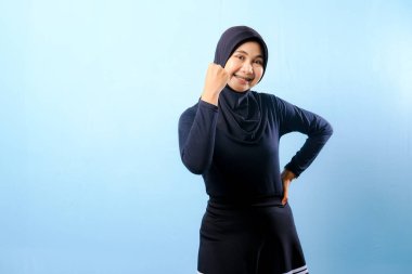 Siyah tesettürlü bir kadın gülümsüyor ve sağ elini kaldırıyor. Mavi bir duvarın önünde duruyor.