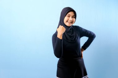 Siyah tesettürlü bir kadın gülümsüyor ve sağ elini kaldırıyor. Mavi bir duvarın önünde duruyor.