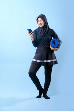 Siyah etekli ve siyah pantolonlu bir kadın mavi yoga minderi tutuyor. Cep telefonuna bakıyor.