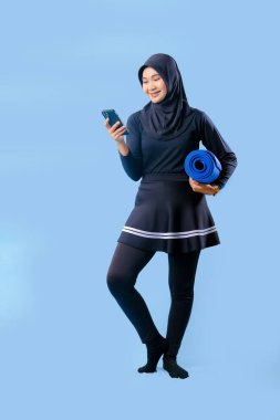 Siyah etekli ve siyah pantolonlu bir kadın mavi yoga minderi tutuyor. Cep telefonuna bakıyor.