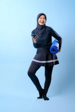 Siyah etekli ve siyah pantolonlu bir kadın mavi yoga minderi tutuyor. Cep telefonuna bakıyor.