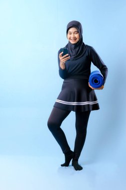 Siyah etekli ve siyah pantolonlu bir kadın mavi yoga minderi tutuyor. Cep telefonuna bakıyor.