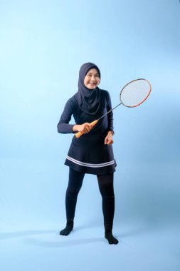 Siyah etekli ve elinde badminton raketi olan siyah pantolonlu bir kadın. Gülümsüyor ve mutlu.