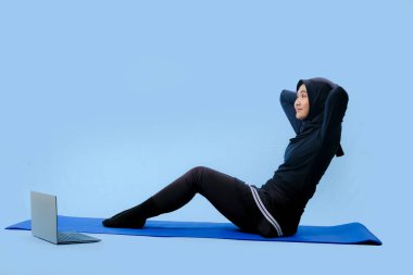 Bir kadın kucağında dizüstü bilgisayarla yoga minderinde oturuyor. Siyah bir bluz ve siyah pantolon giyiyor.