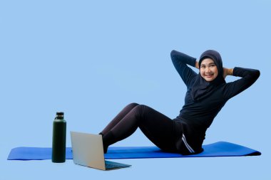 Bir kadın laptopu ve su şişesi olan bir minderde yoga yapıyor. Gülümsüyor ve antrenmanından zevk alıyor.