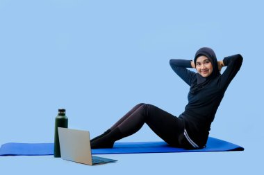 Bir kadın kucağında dizüstü bilgisayarla yoga minderinde oturuyor. Siyah bir bluz ve siyah pantolon giyiyor.