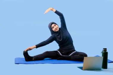 Bir kadın laptopu ve su şişesi olan bir minderde yoga yapıyor. Gülümsüyor ve antrenmanından zevk alıyor.