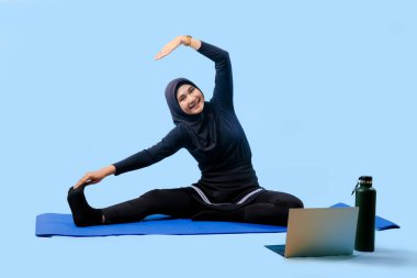 Bir kadın laptopu ve su şişesi olan bir minderde yoga yapıyor. Gülümsüyor ve antrenmanından zevk alıyor.