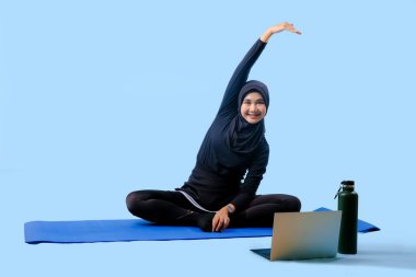 Bir kadın laptopu ve su şişesi olan bir minderde yoga yapıyor. Gülümsüyor ve antrenmanından zevk alıyor.
