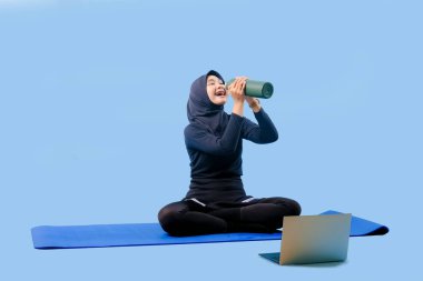 Bir kadın yoga minderinde dizüstü bilgisayarı ve su şişesiyle oturuyor. Gülümsüyor ve eğleniyor.