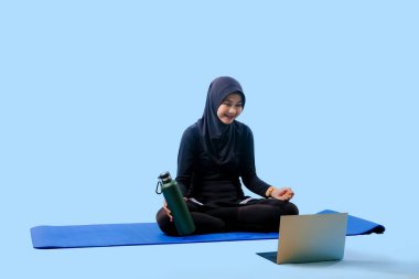 Bir kadın yoga minderinde dizüstü bilgisayarı ve su şişesiyle oturuyor. Gülümsüyor ve eğleniyor.