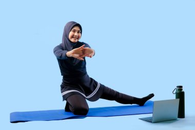 Bir kadın laptopu ve su şişesi olan bir minderde yoga yapıyor. Gülümsüyor ve antrenmanından zevk alıyor.