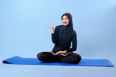 Siyah tesettürlü bir kadın mavi bir paspasın üzerinde oturuyor. Gülümsüyor ve rahatlamış durumda.