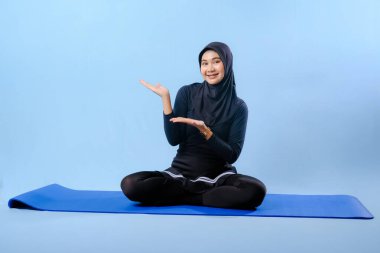 Siyah tesettürlü bir kadın mavi bir paspasın üzerinde oturuyor. Gülümsüyor ve rahatlamış durumda.