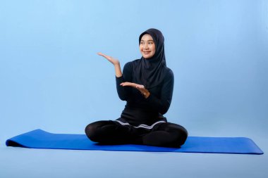 Siyah tesettürlü bir kadın mavi bir paspasın üzerinde oturuyor. Gülümsüyor ve rahatlamış durumda.