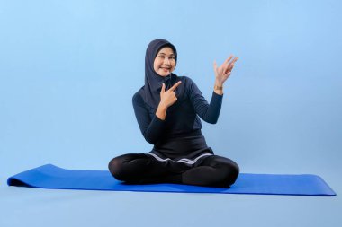 Siyah tesettürlü bir kadın mavi bir paspasın üzerinde oturuyor. Gülümsüyor ve rahatlamış durumda.