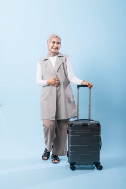 Gri yelek ve beyaz gömlek giyen bir kadın bir bavul tutuyor. Gülümsüyor ve mutlu.