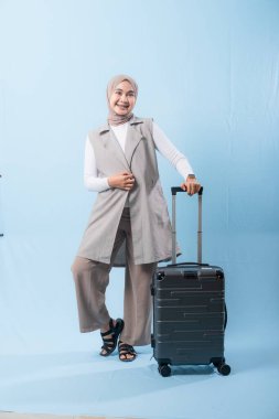 Gri yelek ve beyaz gömlek giyen bir kadın bir bavul tutuyor. Gülümsüyor ve mutlu.