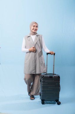 Gri yelek ve beyaz gömlek giyen bir kadın bir bavul tutuyor. Gülümsüyor ve mutlu.