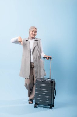 Gri yelek ve beyaz gömlek giyen bir kadın siyah bir bavul tutuyor. Bir şeye işaret ediyor ve gülümsüyor.