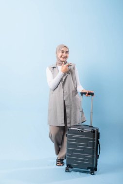 Gri yelek ve beyaz gömlek giyen bir kadın siyah bir bavul tutuyor. Bir şeye işaret ediyor ve gülümsüyor.