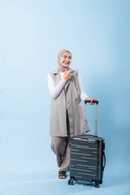 Gri yelek ve beyaz gömlek giyen bir kadın siyah bir bavul tutuyor. Bir şeye işaret ediyor ve gülümsüyor.