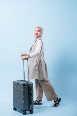 Bir kadın bir bavul tutuyor ve gülümsüyor. Bavul gümüş ve tekerlekleri var. Kadın beyaz bir gömlek ve bir eşarp giyiyor. Seyahat ve macera kavramı