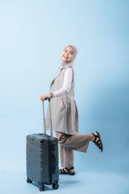 Bir kadın bir bavul tutuyor ve gülümsüyor. Bavul gümüş ve tekerlekleri var. Kadın beyaz bir gömlek ve bir eşarp giyiyor. Seyahat ve macera kavramı