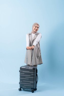 Bir kadın bir bavul tutuyor ve gülümsüyor. Bavul gümüş ve tekerlekleri var. Kadın beyaz bir gömlek ve bir eşarp giyiyor. Seyahat ve macera kavramı