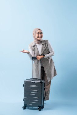 Bir kadın bir bavul tutuyor ve gülümsüyor. Bavul gümüş ve tekerlekleri var. Kadın beyaz bir gömlek ve bir eşarp giyiyor. Seyahat ve macera kavramı