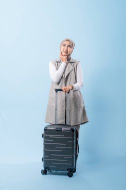 Bir kadın bavulun yanında duruyor, düşünceli görünüyor. Gri bir ceket ve beyaz bir gömlek giyiyor.