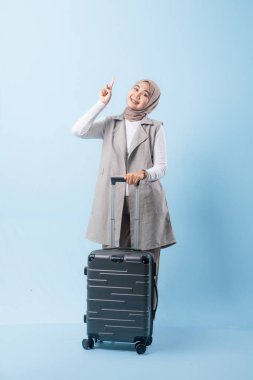 Gri yelek ve beyaz gömlek giyen bir kadın siyah bir bavul tutuyor. Bir şeye işaret ediyor ve gülümsüyor.