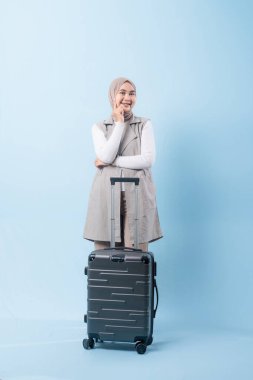 Bir kadın bavulun yanında duruyor, düşünceli görünüyor. Gri bir ceket ve beyaz bir gömlek giyiyor.