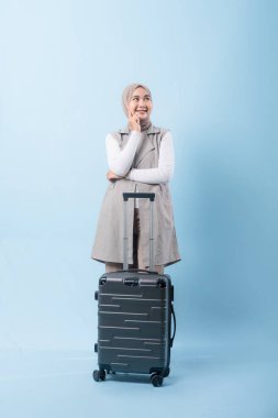 Bir kadın bavulun yanında duruyor, düşünceli görünüyor. Gri bir ceket ve beyaz bir gömlek giyiyor.