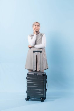 Bir kadın bavulun yanında duruyor, düşünceli görünüyor. Gri bir ceket ve beyaz bir gömlek giyiyor.