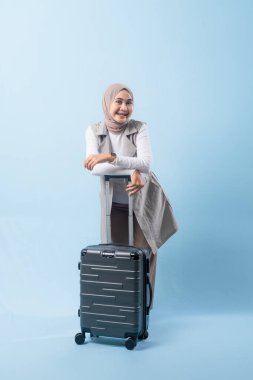 Bir kadın bavulun yanında duruyor, düşünceli görünüyor. Gri bir ceket ve beyaz bir gömlek giyiyor.