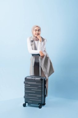 Bir kadın bavulun yanında duruyor, düşünceli görünüyor. Gri bir ceket ve beyaz bir gömlek giyiyor.