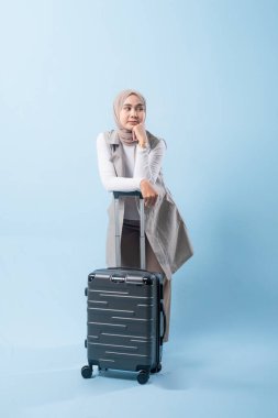 Bir kadın bavulun yanında duruyor, düşünceli görünüyor. Gri bir ceket ve beyaz bir gömlek giyiyor.