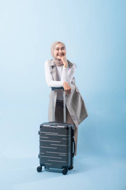 Bir kadın bavuluyla poz veriyor, gülümsüyor ve mutlu görünüyor. Bavul siyah ve tekerlekleri var. Bu da taşımayı kolaylaştırıyor. Kadın beyaz gömlek ve atkı giyiyor.