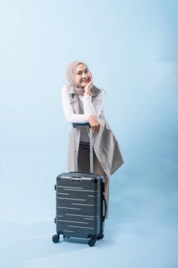 Bir kadın bavuluyla poz veriyor, gülümsüyor ve mutlu görünüyor. Bavul siyah ve tekerlekleri var. Bu da taşımayı kolaylaştırıyor. Kadın beyaz gömlek ve atkı giyiyor.