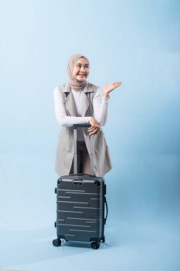 Bir kadın bavuluyla poz veriyor, gülümsüyor ve mutlu görünüyor. Bavul siyah ve tekerlekleri var. Bu da taşımayı kolaylaştırıyor. Kadın beyaz gömlek ve atkı giyiyor.