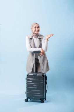 Bir kadın bavuluyla poz veriyor, gülümsüyor ve mutlu görünüyor. Bavul siyah ve tekerlekleri var. Bu da taşımayı kolaylaştırıyor. Kadın beyaz gömlek ve atkı giyiyor.