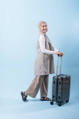 Bir kadın bavuluyla poz veriyor, gülümsüyor ve mutlu görünüyor. Bavul siyah ve tekerlekleri var. Bu da taşımayı kolaylaştırıyor. Kadın beyaz gömlek ve atkı giyiyor.