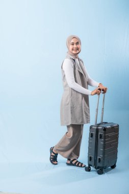 Bir kadın bavuluyla poz veriyor, gülümsüyor ve mutlu görünüyor. Bavul siyah ve tekerlekleri var. Bu da taşımayı kolaylaştırıyor. Kadın beyaz gömlek ve atkı giyiyor.