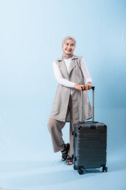 Bir kadın bavuluyla poz veriyor, gülümsüyor ve mutlu görünüyor. Bavul siyah ve tekerlekleri var. Bu da taşımayı kolaylaştırıyor. Kadın beyaz gömlek ve atkı giyiyor.