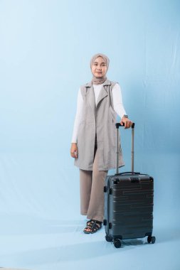 Bir kadın bavuluyla poz veriyor, gülümsüyor ve mutlu görünüyor. Bavul siyah ve tekerlekleri var. Bu da taşımayı kolaylaştırıyor. Kadın beyaz gömlek ve atkı giyiyor.