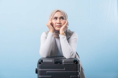 Eşarp takan ve kameraya bakan bir kadın. O bir bavulun üzerinde oturuyor. Derin düşüncelere dalmış.