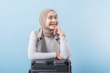 Başörtüsü takan bir kadın bavula oturur. Gülümsüyor ve kameraya bakıyor.