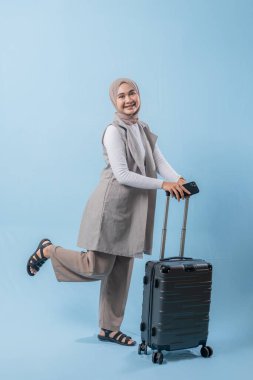 Gri yelek ve ten rengi pantolon giyen bir kadın gülümsüyor ve elinde bavul tutuyor. Çanta siyah ve tekerlekleri var.